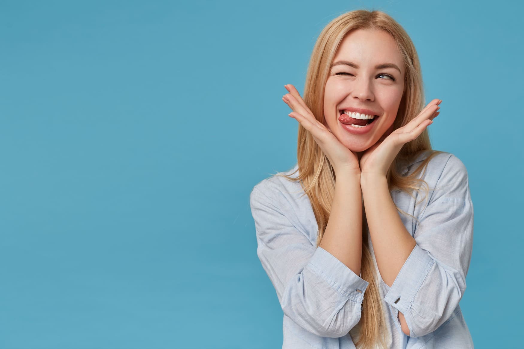 portrait pretty young blonde lady with long hair keeping head raised hands making faces looking aside joyfully with closed eye showinng tongue 1 1